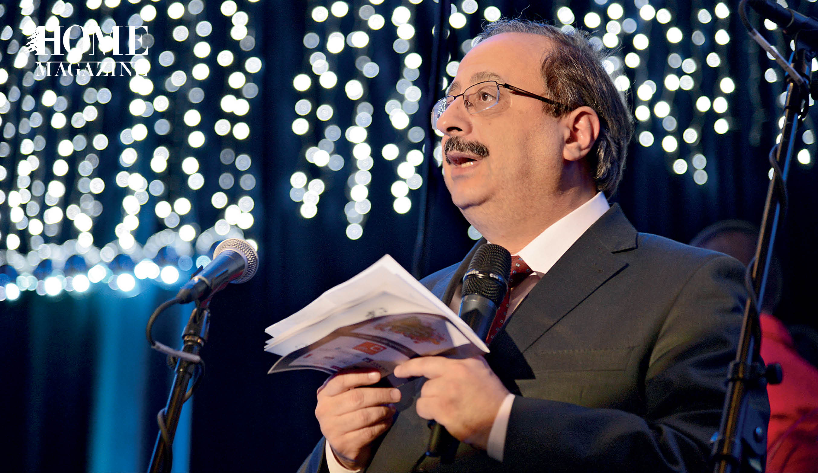 Bald man wearing eyeglasses speaking on a microphone with a notebook in his hands